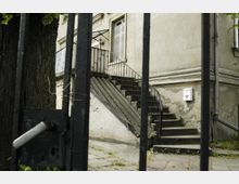 Ansicht eines alten Gebäudes mit bröckelnder Fassade und schmaler Treppe, die zu einer Eingangstür mit Glasvordach führt. Im Vordergrund befinden sich ein schwarzer Metallzaun und ein Baumstamm.