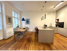 Moderner Wohnraum mit offener Küche und Essbereich. Ein Holztisch mit Stühlen steht vor großen Fenstern, daneben eine Einbauküche mit Kücheninsel, hängenden Lampen und glatten, weißen Schränken; der Boden besteht aus hell glänzendem Holz.