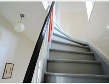 Blick auf eine schmale Treppe mit grauen Stufen und schwarzem Geländer in einem hell gestrichenen Innenraum. Oben an der Wand befindet sich ein rechteckiges Fenster, und an der Decke hängt eine kugelförmige Pendelleuchte.