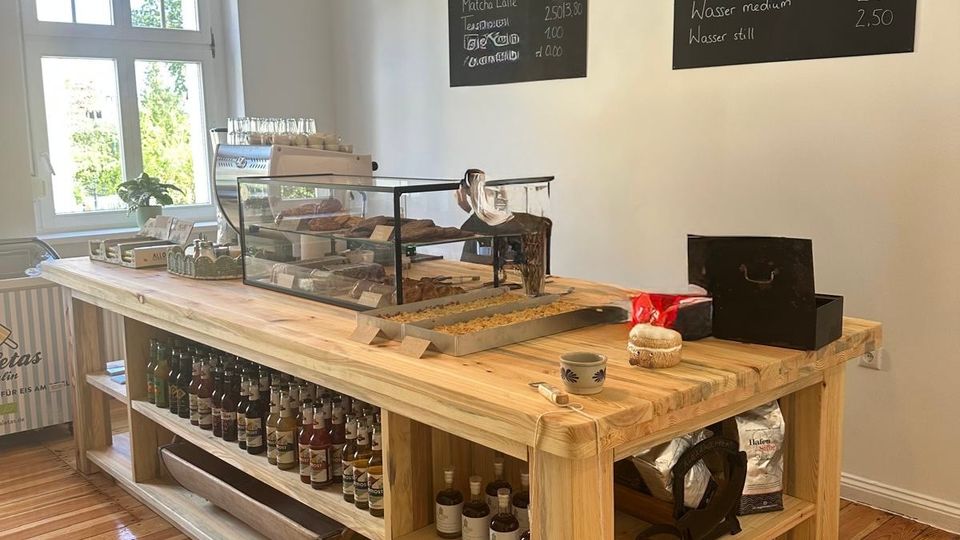 Heller Café-Innenraum mit Holzdielenboden und großem Holzverkaufstisch in der Mitte, darauf eine Glasvitrine und Kaffeezubehör. An der weißen Wand hängen zwei Tafeln mit Getränkekarte, darüber zwei Pendelleuchten; ein Fenster bringt Tageslicht in den Raum.