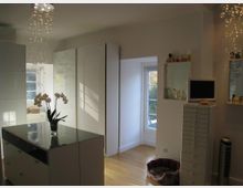 Heller Innenraum mit einer modernen Einrichtung, darunter ein weißer Schrank mit Hochglanzfronten, ein Glastisch mit Orchidee, und ein kleiner Monitor auf einer schmalen Kommode. Ein Fenster mit Blick ins Freie lässt Tageslicht herein, daneben steht ein Haustierbett auf einem Holzboden.