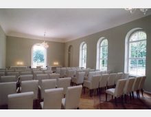 Ein heller Raum mit einer Bestuhlung aus gepolsterten Stühlen, einer Holzfußbodendiele und mehreren hohen, bogenförmigen Fenstern, die Tageslicht hereinlassen und den Blick auf eine grüne Außenlandschaft ermöglichen. An der Stirnwand befinden sich ein großer Bogenfenster, Kerzenleuchter auf Konsolen und ein Kronleuchter an der Decke.