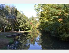 Ein ruhiger Flusslauf, von Bäumen und dichtem Grün flankiert, spiegelt das Ufer und die Vegetation im klaren Wasser wider. Links befindet sich eine kleine, betonierte Plattform und ein Weg, der durch die Bäume führt.