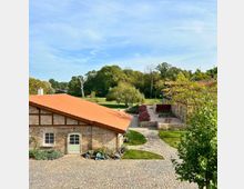 Historischer Gutshof mit Backsteingebäuden und roten Ziegeldächern, umgeben von einem gepflasterten Hof, gepflegten Blumenbeeten und einer grünen Parklandschaft mit Bäumen im Hintergrund. Ein geschwungener Weg führt durch das Gelände.
