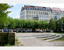 Mehrstöckiges Wohngebäude mit heller Fassade und Schrägdach im Hintergrund, davor eine Reihe dicht bepflanzter Bäume, unter denen geparkte Autos stehen. Im Vordergrund ist eine gepflasterte Fläche mit leicht erhöhten Steintritten.