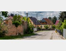 Eine ruhige Wohnstraße mit gepflastertem Gehweg, gesäumt von Einfamilienhäusern mit roten und grauen Satteldächern. Links ist eine alte Ziegelmauer mit Überwucherung zu sehen, rechts befinden sich Gärten und Zäune.