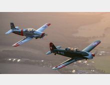 Zwei historische Flugzeuge mit schweizerischen Hoheitszeichen fliegen in Formation über eine ländliche Landschaft bei Sonnenuntergang, mit Feldern und vereinzelten Gebäuden im Hintergrund.