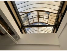 Flur mit einem gewölbten Glasdach, das durch Metallstreben gestützt wird. Im Vordergrund sind weiße Wände und freiliegende Rohre an der Decke sichtbar.