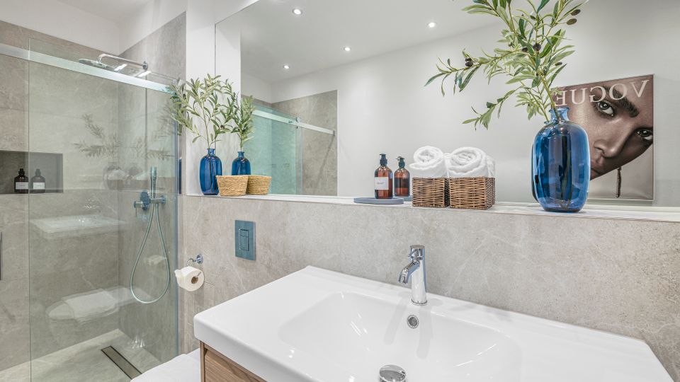 Modernes Badezimmer mit großer, begehbarer Glasdusche, hellgrauen Wandfliesen und einem breiten Waschtisch mit Spiegelwand und Einbaubeleuchtung. Auf der Ablage stehen blaue Glasvasen mit Zweigen, Körbe mit Handtüchern sowie Seifenspender; an der Wand hängt ein dekoratives Bild.