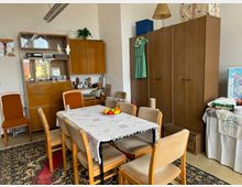 Ein Wohnzimmer im Retro-Stil mit einer Holzessgruppe, bestehend aus einem Tisch mit einer geblümten Tischdecke und sechs Stühlen. Im Hintergrund stehen ein Schrank mit Glasvitrine, Geschirr und Dekorationen sowie ein großer Kleiderschrank; daneben ein Tisch mit einer Karte und gestapelten Textilien. Ein gemusterter Teppich bedeckt den Boden.