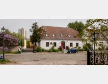 Ein weiß verputztes Gebäude mit Satteldach und mehreren Dachfenstern, umgeben von Bäumen und Sträuchern, liegt an einer Kopfsteinpflasterstraße. Im Hintergrund sind moderne Hochhäuser zu sehen, während eine schmiedeeiserne Zaunspitze im Vordergrund sichtbar ist.