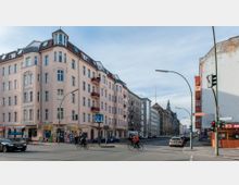 Straßenecke in einer Stadt mit mehrstöckigen Altbauten, teilweise mit Erkern und Balkonen, sowie Geschäften im Erdgeschoss. Im Vordergrund fahren Radfahrer und Autos auf der Kreuzung, während ein U-Bahn-Schild „U“ auf eine U-Bahn-Station hinweist.