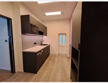 Moderne kleine Küche mit dunklen Schränken und heller Arbeitsplatte, eingebautem Mikrowellenherd und Spüle. Der Raum hat einen hellen Boden, ein schmales Fenster mit weißen Rahmen und indirekte Deckenbeleuchtung.