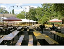 Ein Biergarten mit zahlreichen Holzbänken und Tischen unter freiem Himmel, umgeben von Bäumen und Sträuchern. Im Vordergrund stehen eine Bronzeskulptur und ein Brunnen, während Lichterketten zwischen den Bäumen aufgehängt sind, und große Sonnenschirme sowie ein Zelt im Hintergrund sichtbar sind.