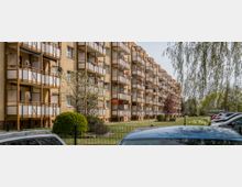 Wohnblock mit mehreren Etagen und Balkonen in beige-gelber Farbe, umgeben von Grünflächen mit Büschen und Bäumen. Im Vordergrund sind geparkte Autos und ein Metallzaun zu sehen.