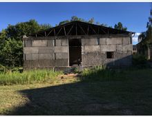 Eine unvollständige Betonhalle mit offenen Wänden und einem Dach aus Holzträgern, umgeben von Gras und Büschen. Im Hintergrund sind Bäume und ein Wohngebäude sichtbar.
