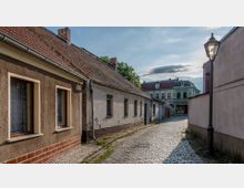 Pflastersteinstraße in einer kleinen Stadt mit einfachen, hell verputzten Häusern mit roten Ziegeldächern. Im Hintergrund ist ein historisches, grünes Gebäude mit dekorativer Fassade zu sehen, rechts steht eine klassische Straßenlaterne.