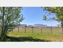 Industriegelände mit großen, weißen Tankbehältern im Hintergrund und einer umzäunten Wiese im Vordergrund. Links und rechts des Bildes stehen einzelne Bäume, die Ausschnittweise sichtbar sind.