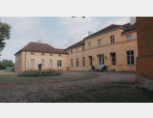 Gelbes, einstöckiges Herrenhaus mit rotem Ziegeldach und symmetrischen Fenstern, umgeben von einem Hof mit Kopfsteinpflaster und einer offenen Rasenfläche. Im Vordergrund befinden sich Blumenbeete und rechts des Eingangs dekorative Pflanzenkübel.