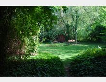 Eine grüne Gartenszene mit einer Rasenfläche, umgeben von dichten Bäumen und Sträuchern. Im Hintergrund steht eine kleine Holzhütte, daneben ein Gartentisch mit Stühlen und ein Heizstrahler.