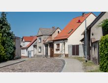 Gepflasterte Dorfstraße mit kleinen Wohnhäusern, teils mit roten Ziegeldächern. Die Fassaden variieren zwischen renoviertem und älterem Zustand, vereinzelt mit Blumenkästen an den Fenstern; am Straßenrand stehen Straßenlaternen und grüne Büsche.