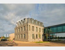 Ein modernes, rechteckiges Gebäude mit grauer Steinfassade und einer Reihe von Dachgauben; rechts davon schließt sich ein verglastet Verbindungsgang an. Im Hintergrund sind ein See und Bäume zu erkennen.