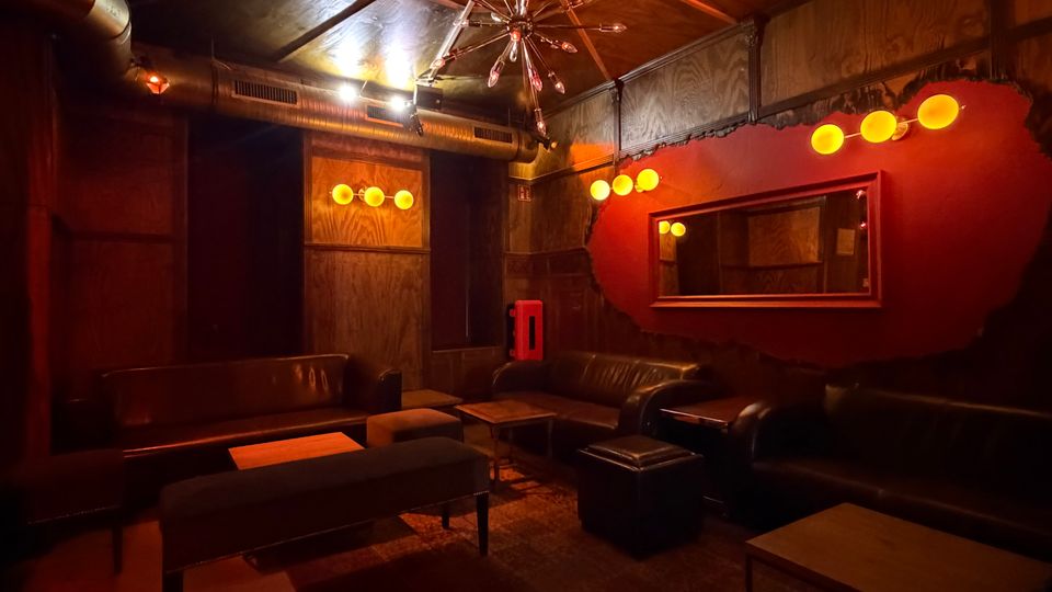 Gedimmter Lounge- oder Barbereich mit dunklen Lederbänken und Sesseln um niedrige Tische, warm beleuchtet von runden Wandlampen und einem zentralen Kronleuchter. Industrielle Details wie sichtbare Lüftungsrohre und Holzvertäfelung prägen den Raum, ergänzt durch eine rote Akzentwand mit großem Spiegel.