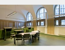 Ein historischer Gerichtssaal mit hohen, gewölbten Decken und großen Fenstern mit Gitterglas. Im Raum befinden sich grüne hölzerne Richterpulte, ein geschwungener Tisch mit grauen Stühlen und holzvertäfelte Wände.