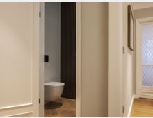 Ein heller Flur mit Holzboden und weißen Wänden führt zu einer halb geöffneten Tür, die einen Blick in ein modernes Badezimmer mit einer wandhängenden Toilette und dunklen Holzakzenten bietet. Im Hintergrund ist eine verglaste Tür mit Rautenmustern sichtbar.