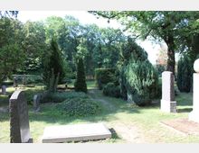 Ein Friedhof mit unterschiedlichen Grabsteinen und dichter Vegetation, darunter hohe Bäume und Sträucher. Im Hintergrund sind ein Holzzaun und ein bewaldetes Gebiet erkennbar.
