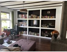 Wohnzimmer mit dunklem Holzboden und weißer Wandregalkonstruktion, die Bücher, Deko und einen Fernseher enthält. Ein großes Fenster mit grünen Vorhängen lässt Tageslicht hinein, während ein rotes Motorrad und ein grauer Kaminofen besondere Akzente setzen.