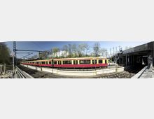 Panoramaansicht eines oberirdischen Bahnsteigs mit einer gelben und roten S-Bahn, die auf den Schienen steht. Im Hintergrund sind Bäume, eine weiße Gebäudefassade und ein klarer Himmel zu sehen; rechts führt ein überdachter Bahnsteig und eine Unterführung.