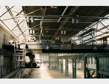 Innenraum einer alten Industriehalle mit hohen Decken und einem Dach aus Glasfenstern, das von Metallträgern gestützt wird. Im Vordergrund ist ein großer Laufkran mit Beschilderung und Maschinenelementen sichtbar, während die Wände aus hellen Ziegeln bestehen.