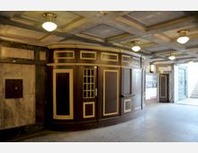 Eingangsbereich einer U-Bahn-Station mit einer abgerundeten Holzkabine in dunkelbrauner Farbe mit goldenen Verzierungen. Darüber befinden sich eine abgehängte Decke mit eingelegten Rechtecken und mehreren runden Deckenleuchten; Treppen führen aus dem Untergrund nach oben.