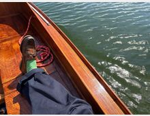 Blick in ein hölzernes Boot mit glänzendem Deck und Bordwand; ein rotes Seil liegt im Boot. Daneben ist die Wasseroberfläche mit leichten Wellen und Sonnenreflexen zu sehen.