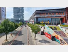 Straßenansicht mit dem "East Side Mall" Einkaufszentrum im rechten Bildbereich, charakterisiert durch eine moderne Glasfassade und großen Schriftzug. Links steht ein mehrstöckiges Bürogebäude, eine Baumreihe säumt die Straße, und im Vordergrund führen bunte Treppen mit Graffiti-Verzierungen zum unteren Bereich der Mall.