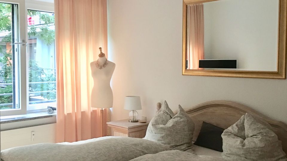 Helles Schlafzimmer mit Doppelbett in Holzoptik, grauer Bettwäsche und mehreren Kissen, daneben ein Nachttisch mit Lampe und ein großer Spiegel an der Wand. Links ein Fenster mit halbtransparenten rosafarbenen Vorhängen und Blick ins Grüne.