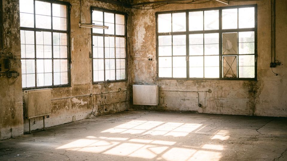 Leerer, heruntergekommener Industrieraum mit großen Sprossenfenstern, abblätterndem Putz und sichtbaren Leitungen an den Wänden. Betonboden mit Sonnenlichtreflexen, Heizkörpern unter den Fenstern und einfachen Leuchtstofflampen an der Decke.