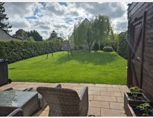 Blick von einer gepflasterten Terrasse mit Rattanmöbeln auf einen großen, gepflegten Rasen im eingezäunten Garten. In der Mitte stehen eine Schaukel und ein großer Baum, umgeben von hohen Hecken sowie einem Holzgartenhaus am rechten Bildrand.