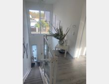 Helles Treppenhaus in einem modernen Wohnhaus mit grauer Treppe, Glasgeländer und Holzfußboden im Obergeschoss. Am Fenster mit Blick auf Nachbarhäuser steht ein kleiner Tisch mit großer Pflanze; unten ist eine Eingangstür mit Glaseinsatz zu sehen.