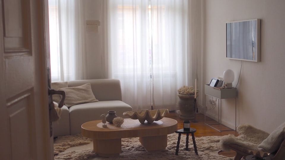 Minimalist living room with tall bay windows covered by sheer white curtains, a low sofa, and a round wooden coffee table on layered shag rugs over wide plank wood flooring. A wall-mounted TV with a small console sits on the right wall beneath a large hanging fabric pendant light.