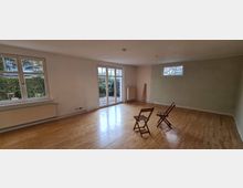 Ein leerer Raum mit Holzboden, weißen Wänden und großen Fenstern, die Tageslicht hereinlassen. Zwei einfache Holzstühle stehen in der Raummitte, und draußen ist ein kleiner Garten mit Hecke sichtbar.