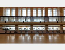 Große, helle Flughafenhalle mit hohen Fenstern, einer verglasten Empore und mehreren Schaltern in einer Reihe im unteren Bereich. Die Architektur ist schlicht mit klaren Linien und viel Glas.