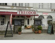 Fassade eines Cafés oder Restaurants mit dem Namen "Theodor" in großen roten Buchstaben auf einem Schild über dem Eingang. Vor dem Gebäude stehen mehrere Menütafeln, Blumentöpfe und draußen sind Sitzgelegenheiten unter einem roten Sonnenschirm zu sehen.