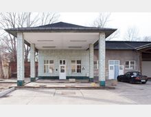 Eine alte Tankstelle mit grünlichen Kacheln an den Säulen und Wänden, einer Überdachung und einer weißen Tür. Im Hintergrund sind ein gemauertes Gebäude und ein schwarzes Auto zu sehen, daneben kahler Baumbestand.