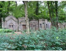 Reihe von historischen Mausoleen mit verzierten Fassaden aus Stein, umgeben von dichtem Wald und üppigem Grün. Im Vordergrund sind Sträucher und Laubbäume zu sehen.
