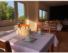 Heller Speiseraum mit einfachen Holztischen und -stühlen, eingedeckt mit weißen Tischdecken, Tassen und Besteck. Große Fenster bieten Blick auf grüne Bäume und lassen Tageslicht in den Raum.