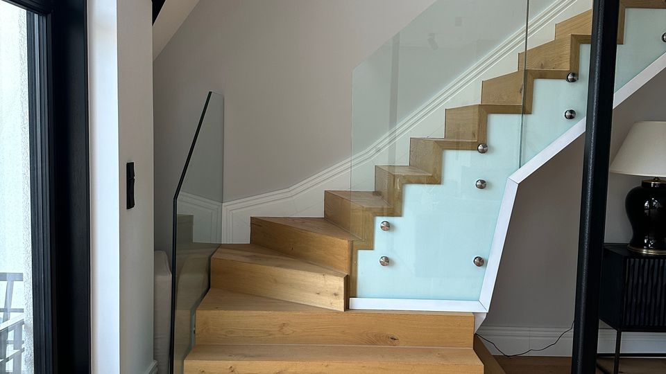 Moderner Innenbereich mit einer Holztreppe, die nach rechts oben führt, und einem seitlichen Glasgeländer mit Metallhaltern. Links befindet sich eine große Glastür oder Fensterfront, darunter ein Holzfußboden; rechts ist ein schwarzer Stützpfosten zu sehen.