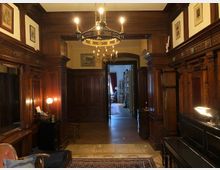 Holzvertäfelter Wohnraum mit antiken Möbeln, einem Kamin und einem Kronleuchter an der Decke. Durch eine offene Tür ist ein angrenzender Raum mit Bücherregalen und Tageslicht zu sehen.