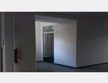 Ein minimalistischer Innenraumflur mit beigen Wänden und grauem Boden. Im Vordergrund ist ein Feuerlöscher an der Wand befestigt, im Hintergrund führt eine offene Tür zu einem weiteren Raum mit einem Heizkörper und verglastem Oberlicht.